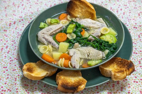 Homemade chicken wing soup with carrots, chayote, potato, spinach and small pasta shells in a clear broth, served in a green ceramic bowl with toasted bread on a floral tablecloth.