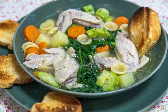 Homemade chicken wing soup with carrots, chayote, potato, spinach and small pasta shells in a clear broth, served in a green ceramic bowl with toasted bread on a floral tablecloth.