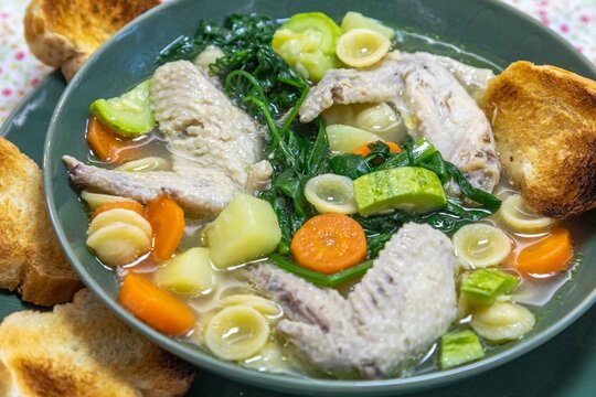 Homemade chicken wing soup with carrots, chayote, potato, spinach and small pasta shells in a clear broth, served in a green ceramic bowl with toasted bread on a floral tablecloth.