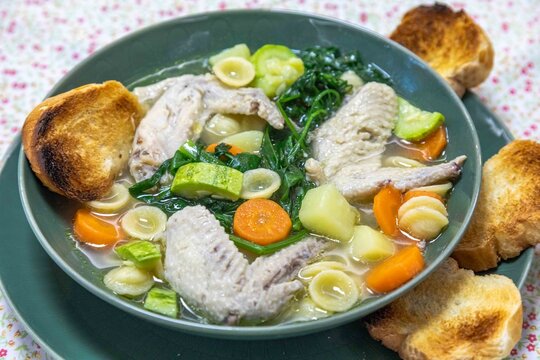 Homemade chicken wing soup with carrots, chayote, potato, spinach and small pasta shells in a clear broth, served in a green ceramic bowl with toasted bread on a floral tablecloth.