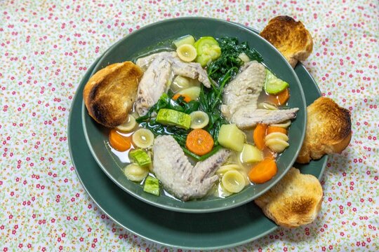 Homemade chicken wing soup with carrots, chayote, potato, spinach and small pasta shells in a clear broth, served in a green ceramic bowl with toasted bread on a floral tablecloth.