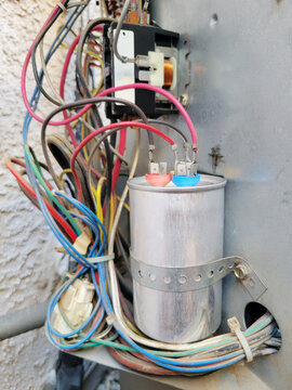 A brand new shiny dual motor run capacitor connected to multiple colorful electrical wires inside an outdoor HVAC system compressor housing