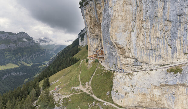 Aerial view of Aescher Guesthouse built into the cliffside with a narrow mountain path, lush green slopes, and dramatic rock formations in Schwende-Rute, Appenzell Innerrhoden, Switzerland.