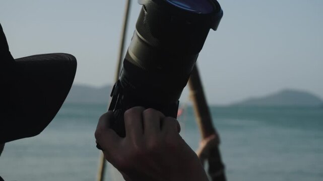 Closeup back view of male photographer taking pictures of a bikini model posing on the beach against sea and mountains background. Working process and back stage of fashion photo session on the coast.