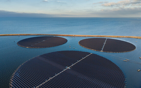 Aerial view of circular floating solar panel arrays installed on a calm body of water with a long dike in the background in The Netherlands.
