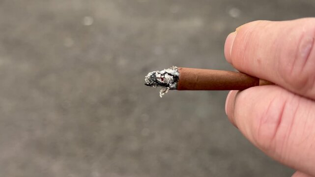 Close up shot of a man holding a burning brown cigarette with ash between his fingers outdoors. Smoking habit concept, tobacco product use, male hand close-up, unhealthy lifestyle choice.