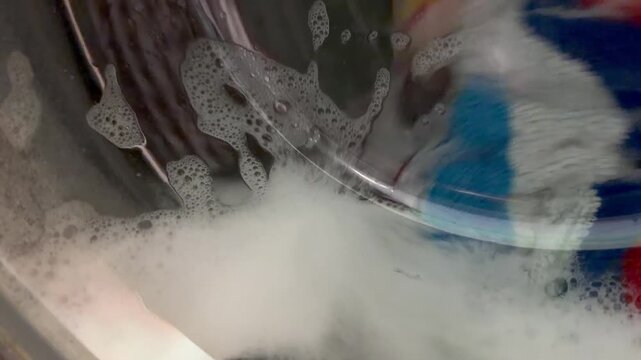Colorful towels are washed with white soap foam inside a washing machine. Domestic laundry process, home appliance cleaning, bright textile wash, everyday housework routine.
