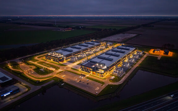 Aerial view of a large illuminated data center facility at night, surrounded by dark agricultural fields, roads, and a small pond in The Netherlands.