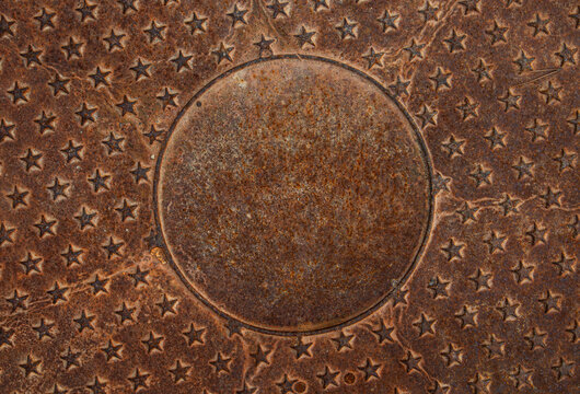 Close view of a rusted cast iron manhole cover with a star pattern and circular center in an urban setting