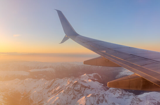 Stunning aerial view of mountain range at sunrise or sunset from airplane window, highlighting dramatic sky, clouds, and golden light on snow-capped peaks.