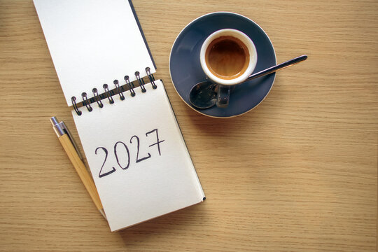 New Year 2027 resolutions on the desk. A notepad with handwritten text 2027 and a cup of espresso on a wooden table. Goals, plan, checklist concept, grainy film texture, high quality photo