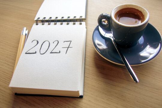 New Year 2027 resolutions on the desk. A notepad with handwritten text 2027 and a cup of espresso on a wooden table. Goals, plan, checklist concept, grainy film texture, high quality photo