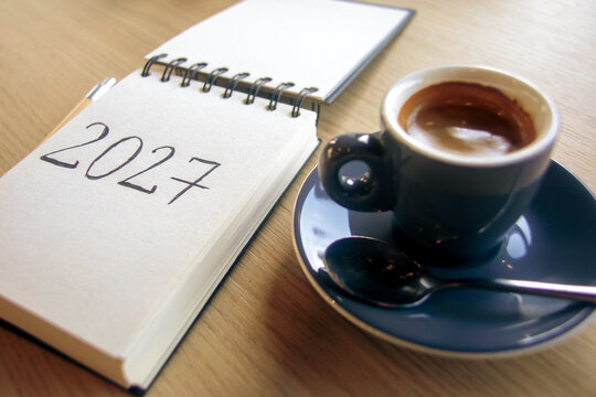 New Year 2027 resolutions on the desk. A notepad with handwritten text 2027 and a cup of espresso on a wooden table. Goals, plan, checklist concept, grainy film texture, high quality photo