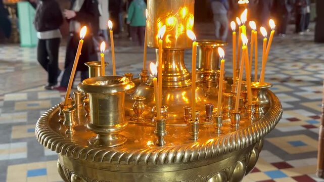 Close up shot of thin beeswax candles burning on  golden brass candlestick inside a church as people walk in the background. Russian orthodox cathedral interior, religious prayer devotion ritual.