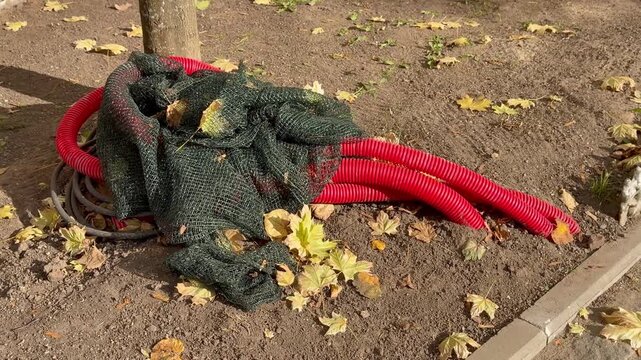 Flexible corrugated red plastic pipes and dark protective mesh lie on the ground under a tree during autumn construction works outdoors. Urban utility installation, street renovation service.
