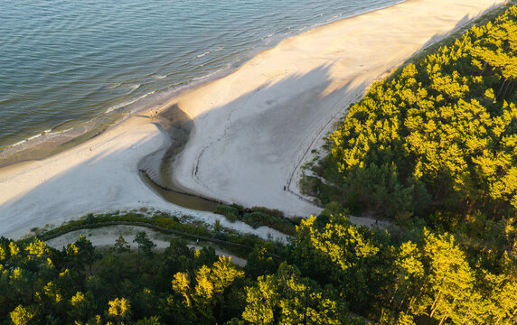 Aerial view of the sandy beach and coastline featuring a small stream flowing into the Baltic Sea, bordered by pine forests in Karwia, Pomeranian Voivodeship, Poland.