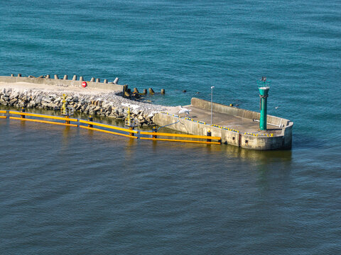 Aerial view of a harbor breakwater with a green lighthouse and yellow safety barriers extending into the sea in Leba, Pomeranian Voivodeship, Poland.