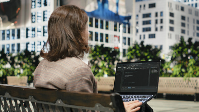Female programmer writes code on a laptop from a park bench and uses a coding interface while working remotely in an outdoor setting.