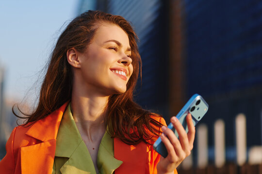 Crypto trader, joyful young woman in profile with open happy smile holds smartphone in sunny city street. Concept of successful mobile trading and digital investment lifestyle.
