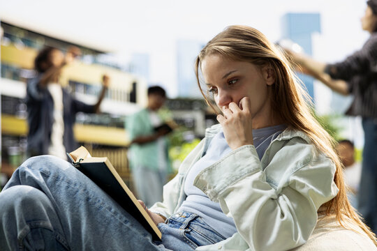 Young student reads an open book on a beanbag outdoors, focusing on solo study, memorizing notes and exam preparation during a modern campus learning session with peers and study materials nearby.