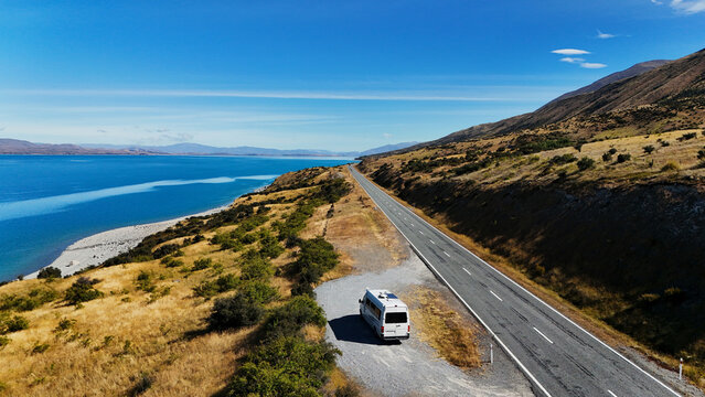 Aerial view of a campervan parked at Peter's Lookout overlooking the turquoise waters of Lake Pukaki with a winding road and golden hills in Ben Ohau, Canterbury Region, New Zealand.