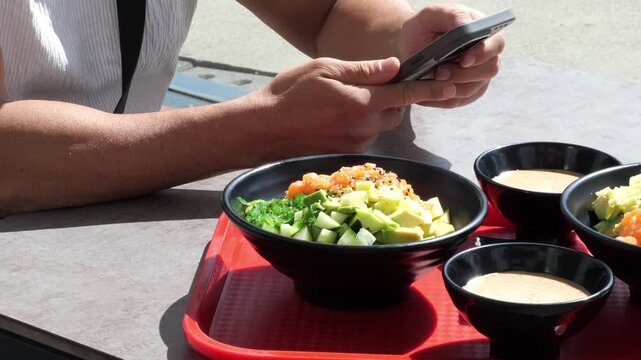 Person Holding Smartphone next to Fresh Hawaiian Salmon Poke Bowls with Avocado Seaweed and Dressing Sauces at Outdoor Restaurant Table Food Concept