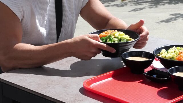 Young Athletic Man Eating Fresh Hawaiian Salmon Poke Bowl with Fork at Outdoor Restaurant Terrace Table Healthy Fast Casual Dining Concept
