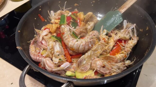 Close-up view of whole mantis shrimp, bell peppers, zucchini, and garlic being stir-fried with a silicone spatula in a large wok pan on a stove.   
