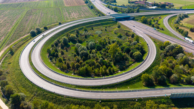 Aerial view of A58 Motorway interchange with a circular ramp surrounded by lush green fields and trees in the rural landscape of Truccazzano, Lombardy, Italy.