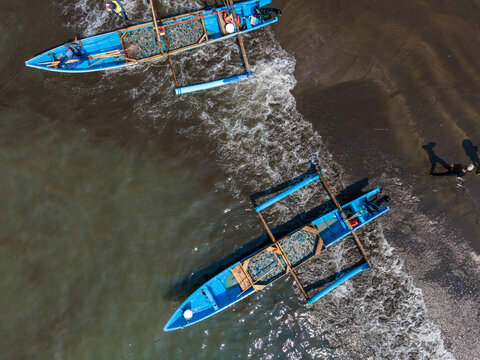 Aerial view of two traditional blue outrigger boats resting on the sandy shore with gentle waves washing over the beach in Cilacap, Central Java, Indonesia.