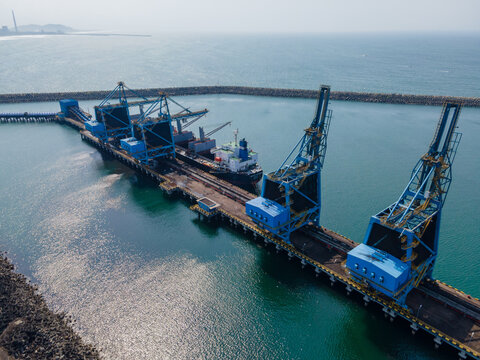 Aerial view of large blue industrial cranes loading a cargo ship at a port facility with a breakwater extending into the sea in Cilacap, Central Java, Indonesia.