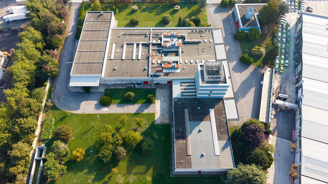 Aerial view of the Cinemeccanica industrial facility featuring a large flat roof with ventilation units, office section, green lawns, and parking areas in Caleppio, Lombardia, Italy.