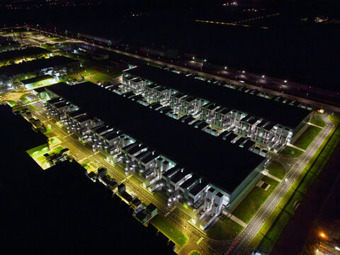 Aerial view of a large data center facility illuminated at night, featuring rows of server buildings, cooling infrastructure, and surrounding access roads in the Netherlands.