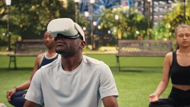 Man wears a VR headset and sits cross-legged on yoga mat in city park, starting outdoor meditation with virtual reality in lotus position.