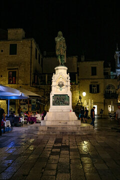 View of the bronze statue of Ivan Vucetic on a stone pedestal in the main square at night, with historic buildings and outdoor seating in Hvar, Split-Dalmatia County, Croatia.