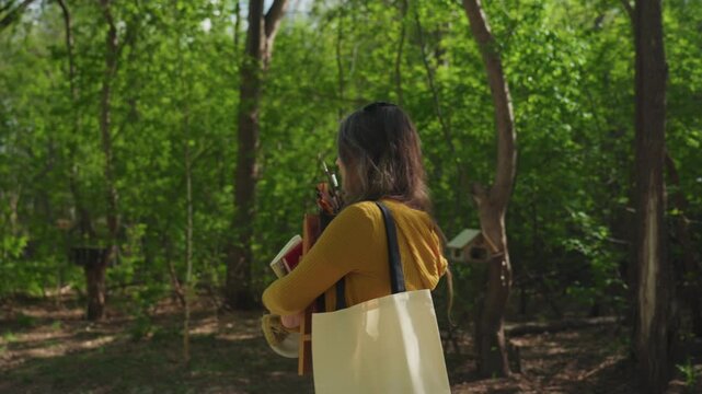 Woman carries painting supplies through forest path. Woman is seen from behind with long hair in loose waves wearing mustard yellow sweater over black top and carrying off white tote bag