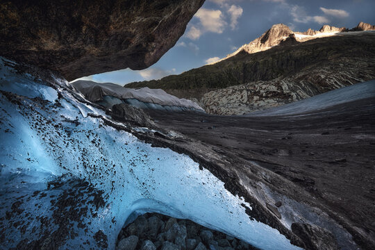 View of the vibrant blue ice of the Rhone Glacier protected by thermal blankets against a backdrop of rugged mountains and rocky terrain in Obergoms, Valais, Switzerland.