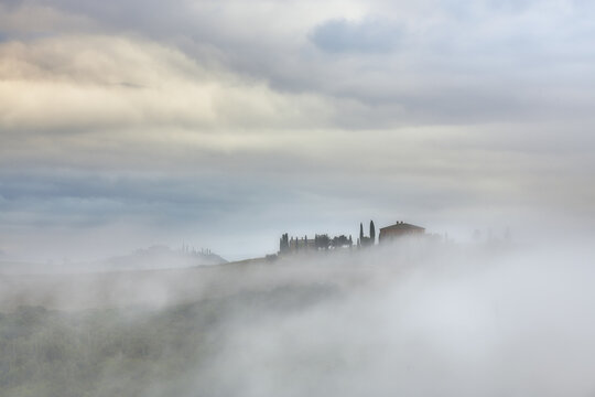 View of a traditional farmhouse surrounded by cypress trees emerging from a thick morning fog in the rolling hills of the countryside, Tuscany, Italy.