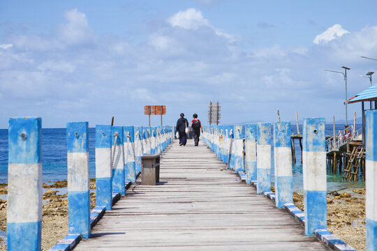 View of two people walking along a wooden pier with blue and white painted posts extending into the sea under a blue sky in Bali, Indonesia.