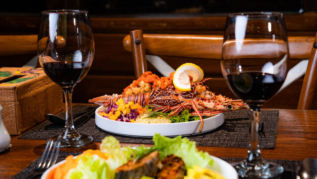 View of a gourmet lobster dish served with lemon, corn, and fresh salad on a wooden restaurant table with two glasses of red wine in Indonesia.