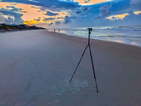 View of a camera on a tripod on the sandy beach of Topsail Island at sunset, with people walking in the distance and dramatic clouds in North Topsail Beach, North Carolina, United States.