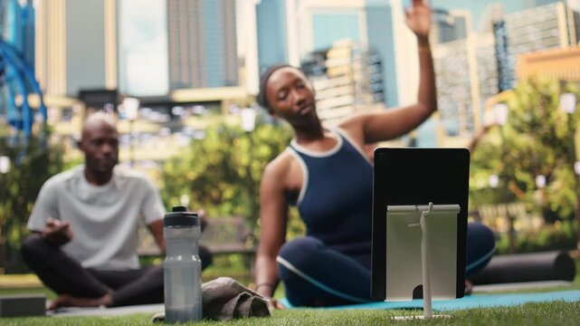 Woman instructor does a side stretch exercise in front of tablet with participants, following a digital yoga tutorial in public park while stretching together on a mat for wellness.