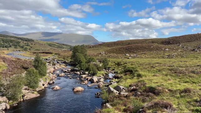 A peaceful river flows through a green meadow surrounded by hills and a cloudy sky, Ullapool, Highlands, Scotland, United Kingdom