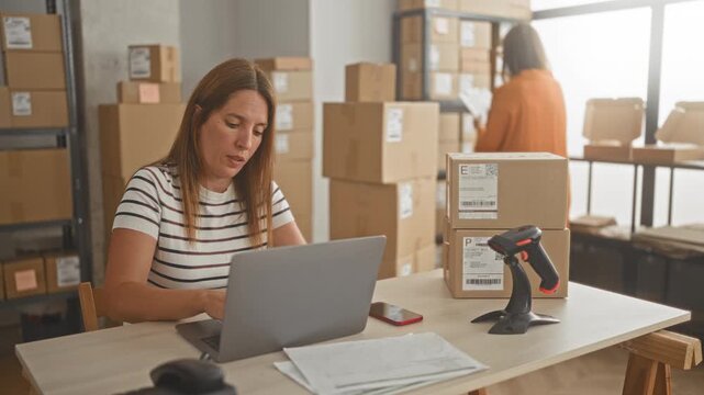 Woman typing on laptop with barcode scanner, stacked cardboard parcels and smartphone in building packing area; teamwork productivity shipping.