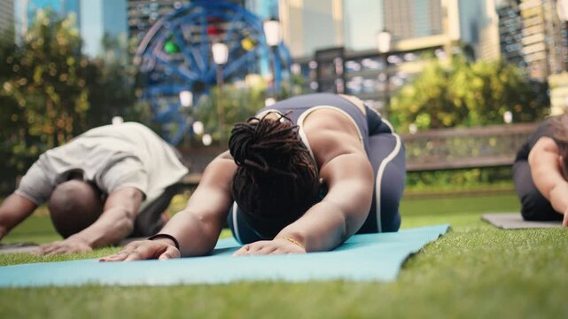Yoga participants extend arms forward in child pose on exercise mats, practicing a bent-over stretch during outdoor class in city park and supporting recovery workout and muscle relief.