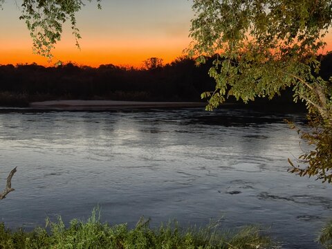 View of the Okavango Delta at sunset with calm river waters reflecting the orange sky and lush riverside vegetation in North-West District, Botswana.
