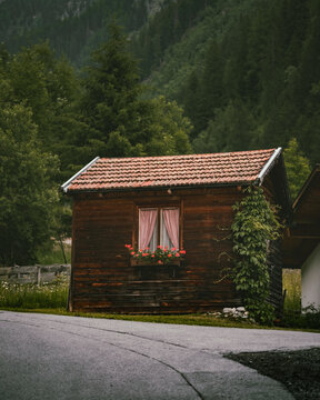 View of a rustic wooden cabin with a tiled roof and a window box filled with red flowers, situated along a quiet road in the alpine landscape of Kaunertal, Tyrol, Austria.