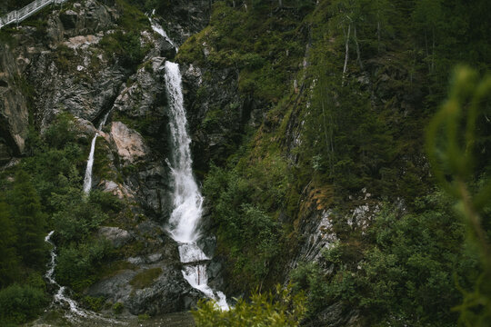 View of the waterfall cascading down rocky cliffs surrounded by lush green forest and pine trees in Kaunertal, Tyrol, Austria.