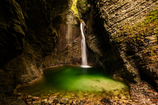 View of the emerald green pool and waterfall of Kozjak in a narrow rocky gorge surrounded by mossy stone walls in Slovenia.