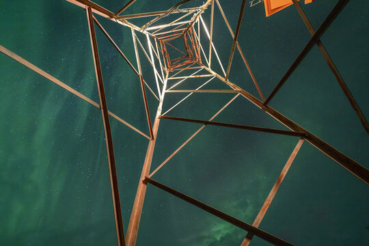 View of a metal transmission tower silhouetted against the vibrant green Aurora Borealis in the night sky in Iceland.